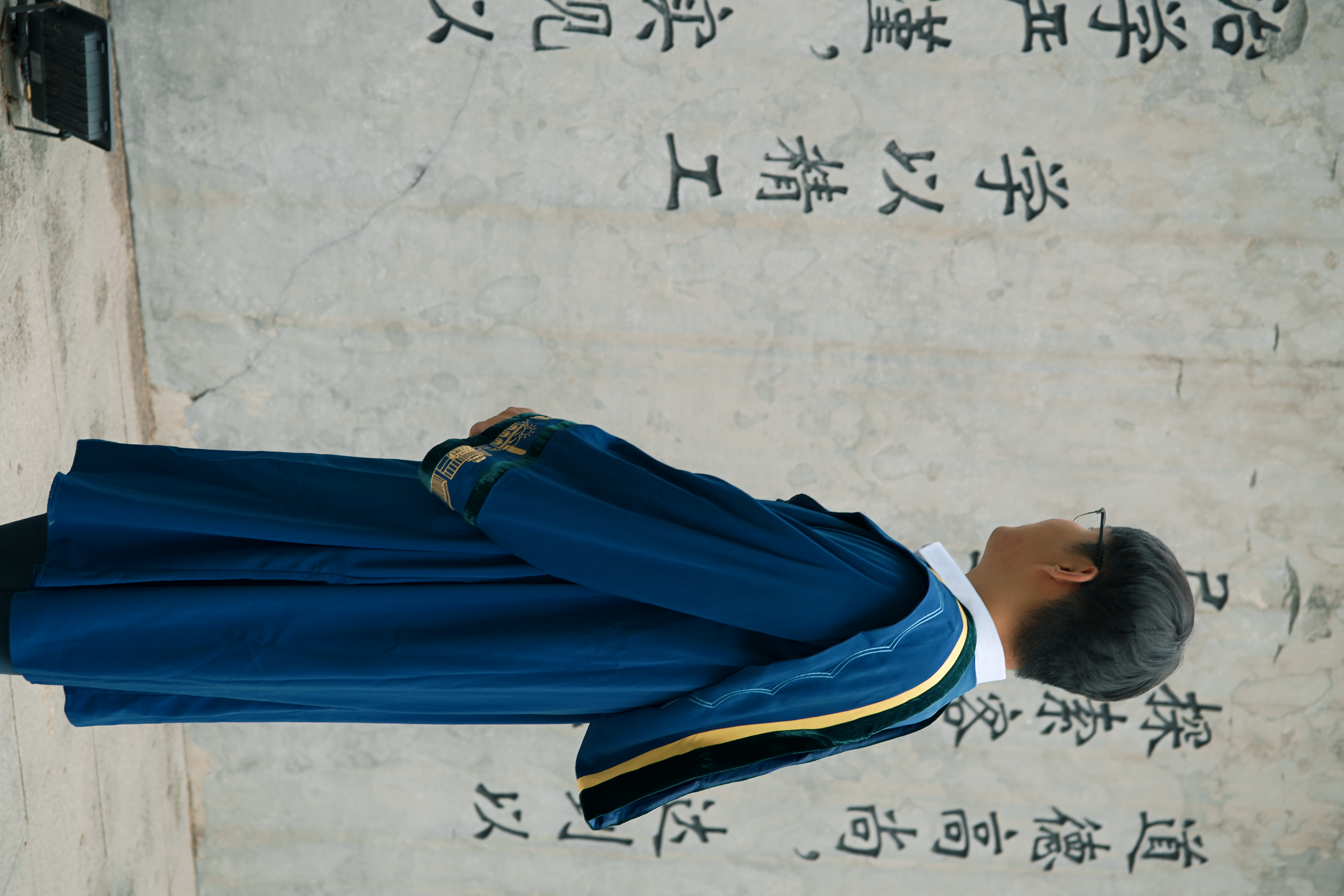 一位身着蓝色学位服的毕业生站在林荫道路旁
